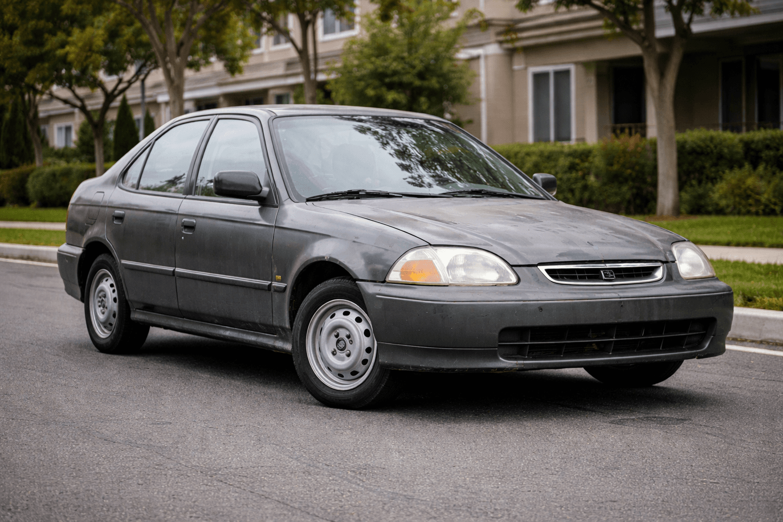 Weathered dark gray Honda Civic sedan parked on a residential street with houses and trees.